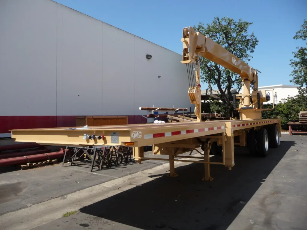 Yellow QMC trailer-mounted crane with extended boom on flatbed, highlighting specialized boom truck crane models built for transporting and lifting heavy loads.